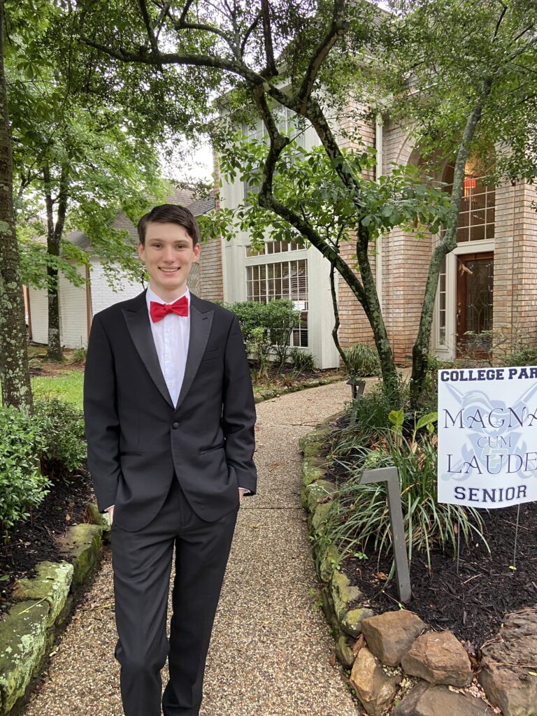 A photograph of Jordyn's friend Murdo. He has brown short hair, is smiling and wearing a black suit with a red bow-tie. Behind him is a sand-coloured building and green trees. 