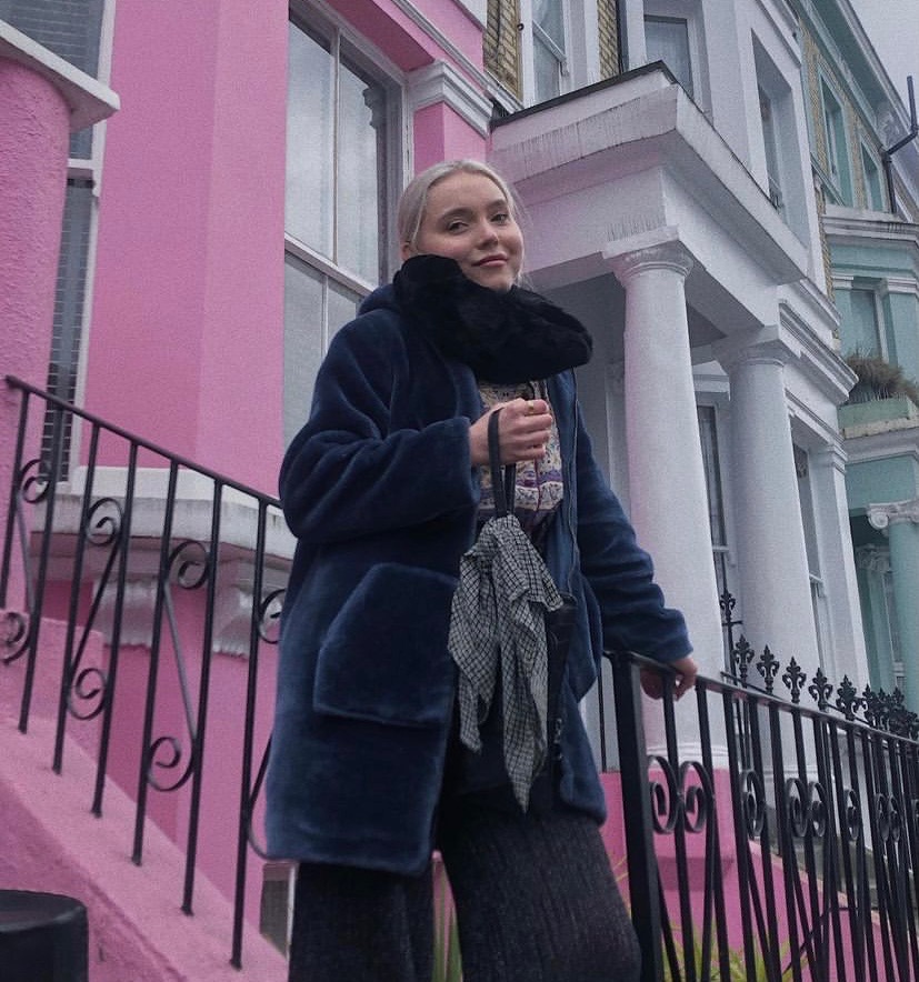 A photograph of Jordyn's friend Gro. She has blond hair, is smiling and standing on the steps outside a pink building.