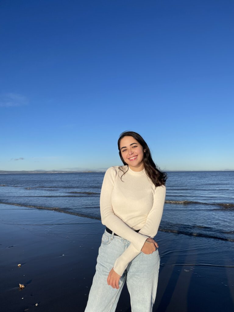 A photograph of Kenza Darrak. It displays Kenza standing on the beach with sea behind her. Kenza has black hair and is wearing a white sweater and light blue jeans. She is smiling gently.
