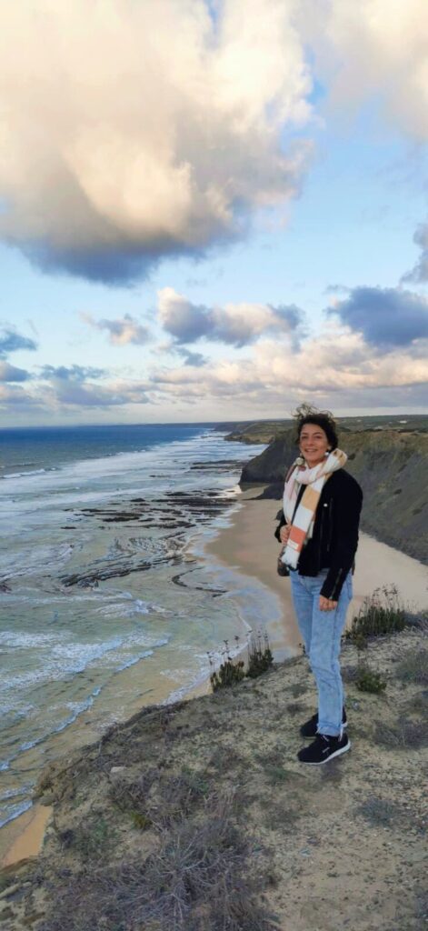 A photograph of Jordyn's friend Olivia. She is standing besides a gray cliff near the sea. She is wearing a scarf, warm sweater and jeans. It looks windy. 