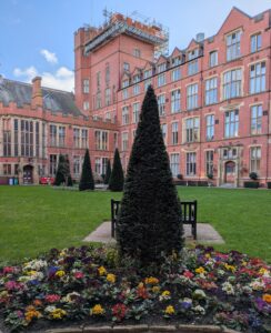 External view of red-brick university building in the background with trees and flowers in the foreground.