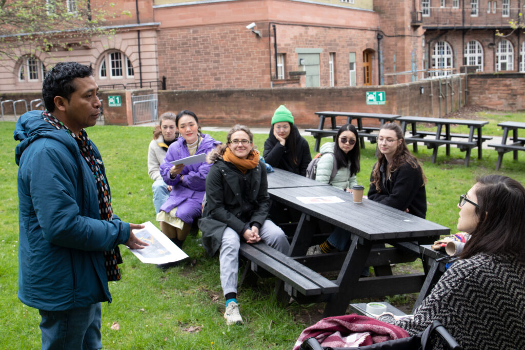 Colour photograph showing artist Antonio Pichilla as he is speaking to a group of students and faculty seated around picnic tables on the ECA campus courtyard. He is seen from the side, with short hair, wearing a blue jacket over a shirt with colourful geometric designs, bright green grass in the background.