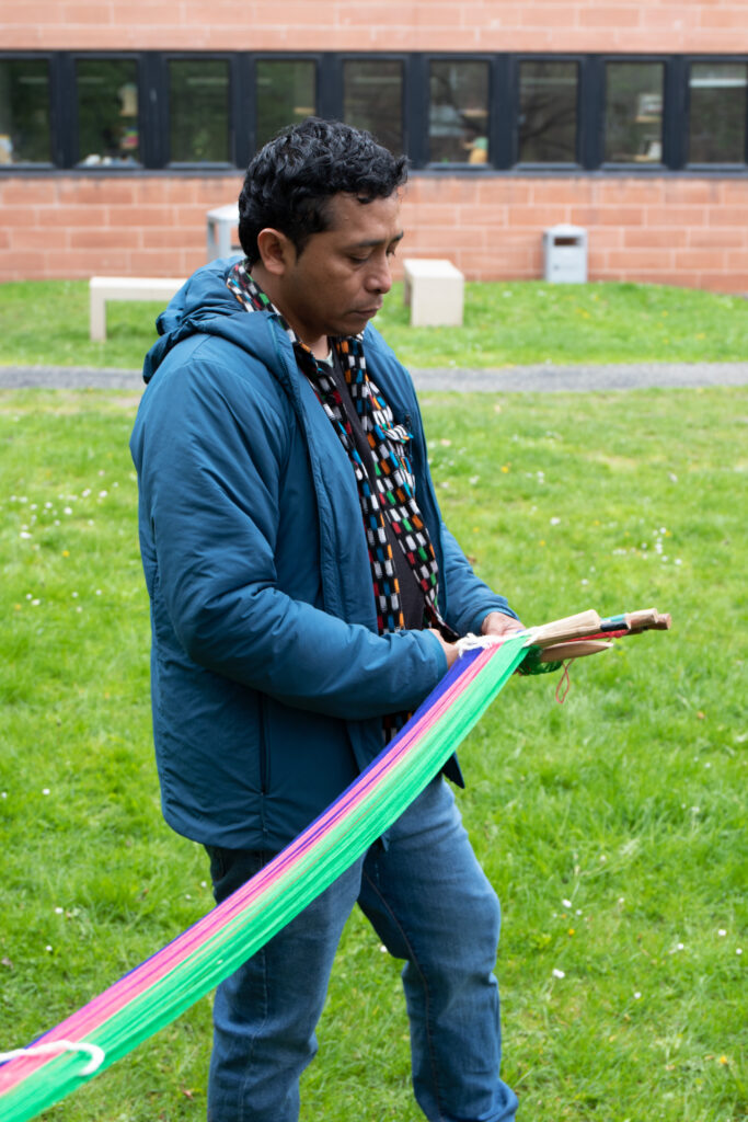 Colour photograph showing artist Antonio Pichilla is holding a backstrap loom, with threads in green, pink and blue. He is seen from the side, with short hair, wearing a blue jacket over a shirt with colourful geometric designs, bright green grass in the background.