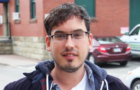 Stuart Dunmore wears a dark blue hooded sweatshirt and looks at the camera in front of a red brick building in Antigonish, Nova Scotia.