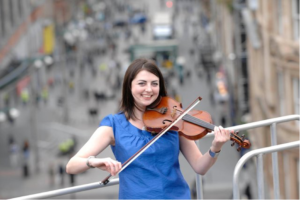 Lori Watson plays the fiddle on a rooftop wearing a blue dress infron of a busy city street.