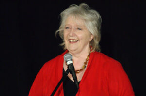 Nancy Nicolson stands at a microphone smiling. She is wearing a red cardigan and has silvery hair against a black backdrop.