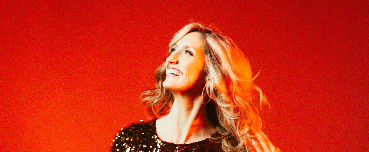 Joy Dunlop smiles and looks to her right wearing a bronze sequinned top, with blond curls blowing in a light breeze against an orange-red background.