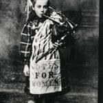 Young Bessie Watson stands wearing a kilt, playing the bagpipes, with a"Votes for Women" sash.