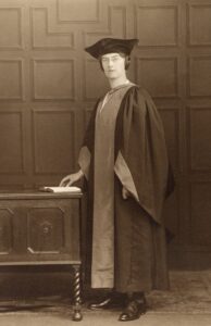 Annie Hutton Numbers wears a graduation robe and hat, standing in front of a wooden wall.