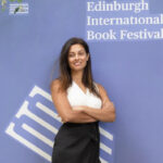 Devi stands with arms crossed in front of a purple "Edinburgh International Book Festival" background