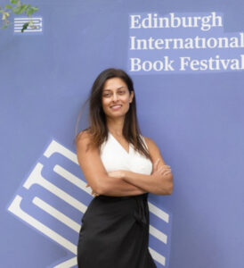 Devi stands with arms crossed in front of a purple "Edinburgh International Book Festival" background