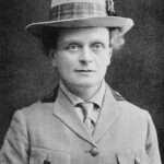 Elsie Inglis looks at the viewer. She is wearing a buttoned jacket, tie, and hat with plaid ribbon.