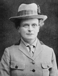 Elsie Inglis looks at the viewer. She is wearing a buttoned jacket, tie, and hat with plaid ribbon.