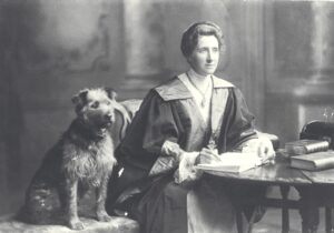 Jessie sits at her desk in graduation robes, writing in a book. A scruffy dog sits next to her.