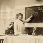 Gertrude Hertz wears a lab coat and points to a scientific figure on a black board.