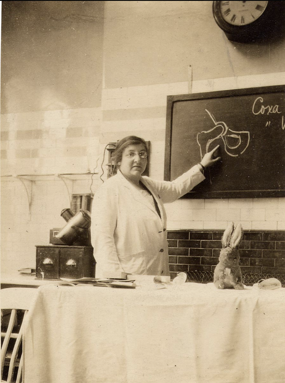 Gertrude Hertz wears a lab coat and points to a scientific figure on a black board.