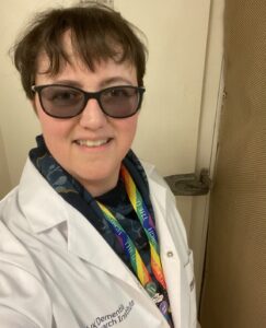 Tara wears her lab coat and lanyard, smiling into camera