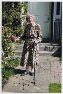 Brenda Moon stands with a cane in her garden, touching a rose, wearing a floral dress and smiling.