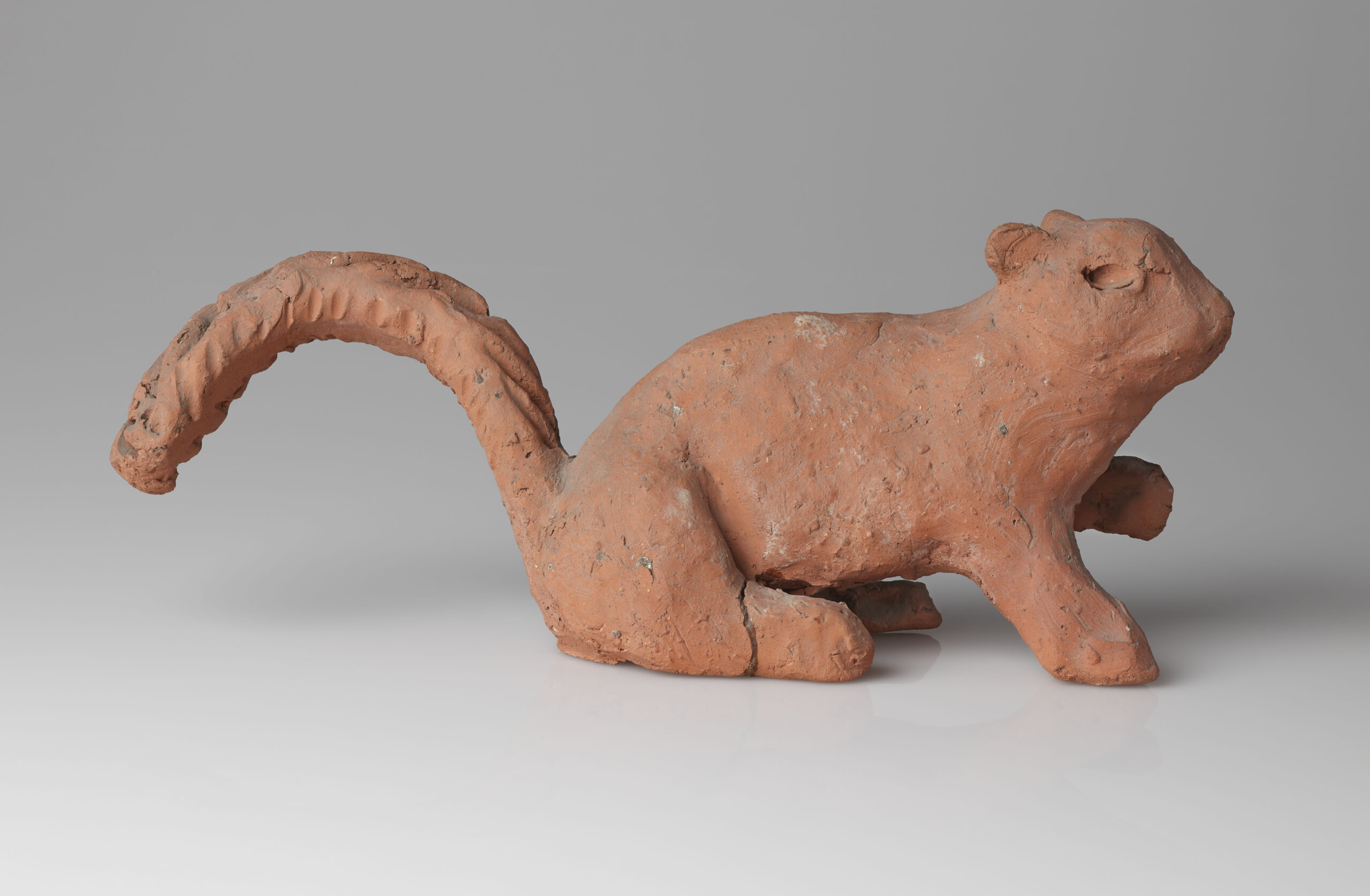 Small unglazed ceramic sculpture of a squirrel. The Squirrel has been decorated with a rust-red slip and has its right paw raised in the air.