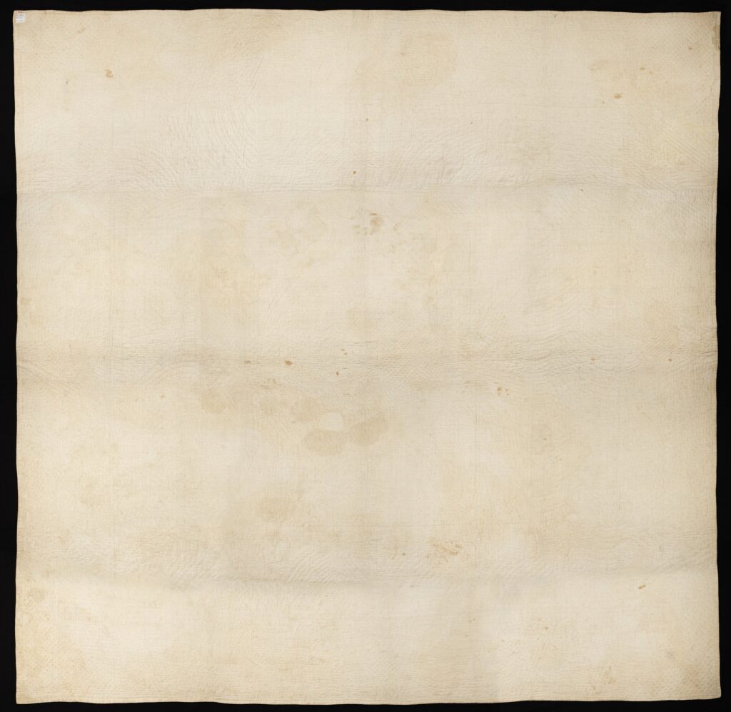 Back of large, square patchwork quilt. Intricate stitching from the construction of the quilt can be seen on a pale cream fabric.