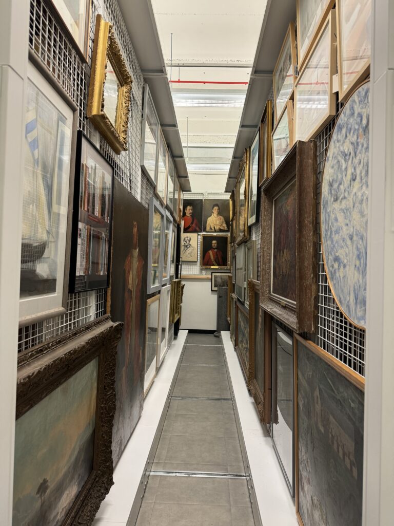 View of corridor with metal grids of shelves on either side. Each grid has a number of framed artworks hanging on them. At the far end of the corridor , portraits can be seen.