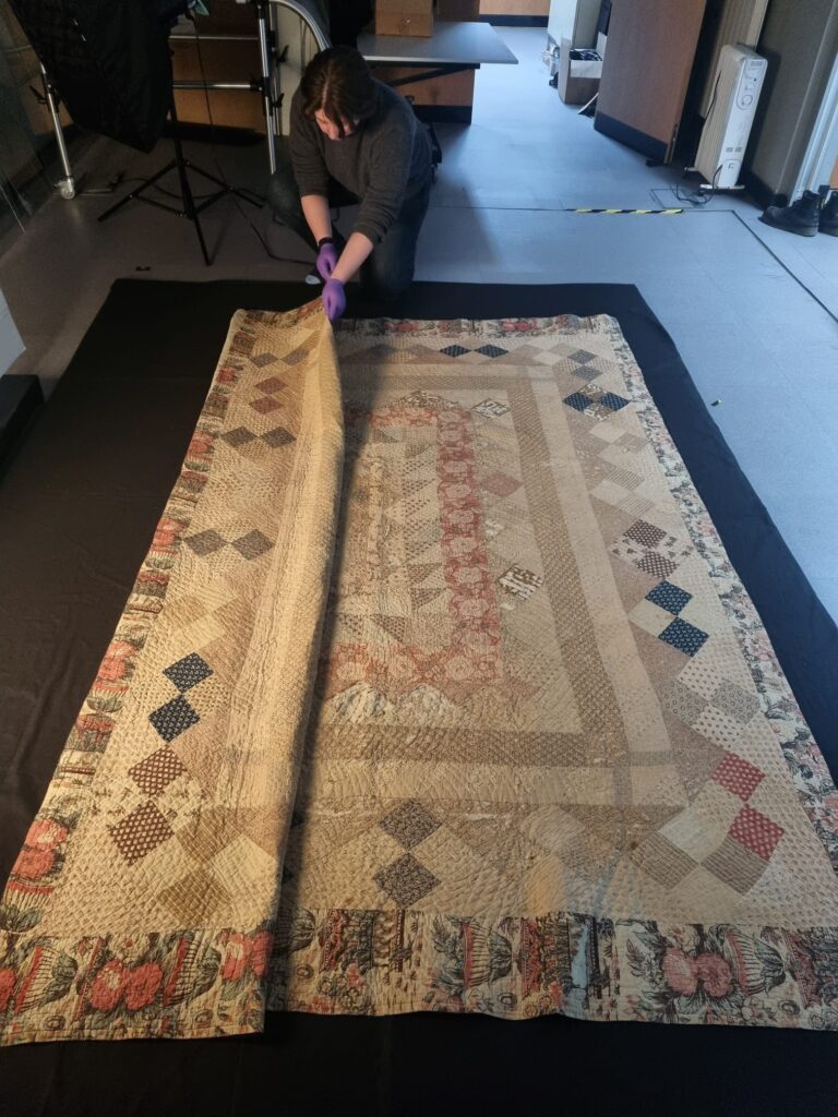 Image of Gaby Cortes handling the front of the quilt. The quilt is laid flat on the floor, lying on black foam. She is crouched, folding the large quilt in sections.