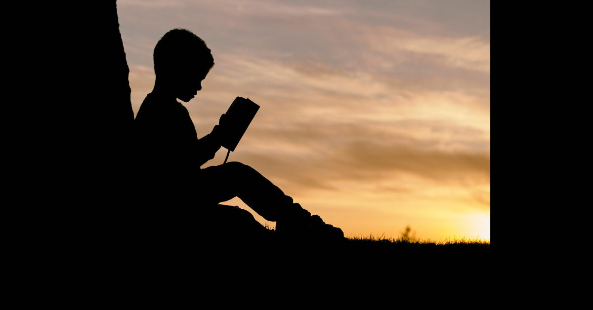 The silhoutte of a child leaning against a tree, reading.