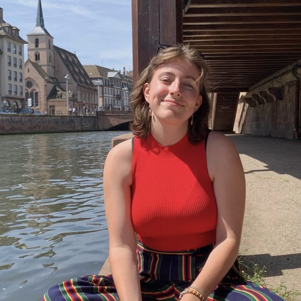 A profile image of Zita. She is sitting on a sunny bridge by a waterway, and smiling. A line of buildings and a church can be seen in the background.