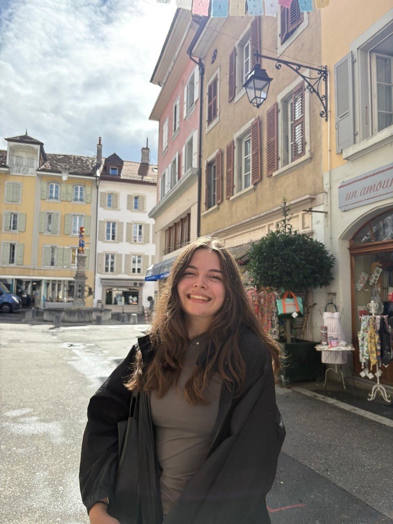 A profile image of Alicia. She is stood on a sunny street full of colourful buildings and shops, with a stone fountain behind her.