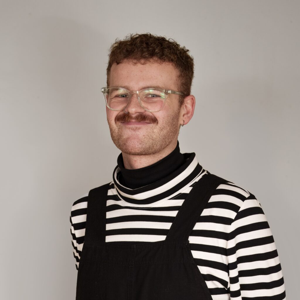 A profile image of Euan. They are smiling and wearing a black and white striped shirt, with black dungarees.