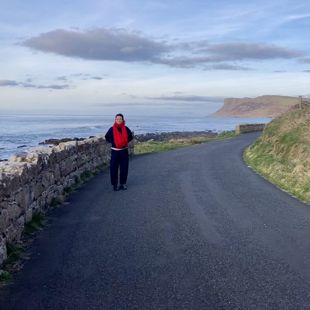 A profile image of Niamh. She is standing on a rural road with the ocean to her back, and a cliff is framed in the distance.