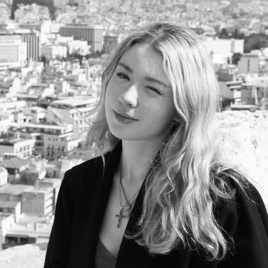 A monochrome profile image of Regan. She is winking at the camera, with an out of focus cityscape behind her.