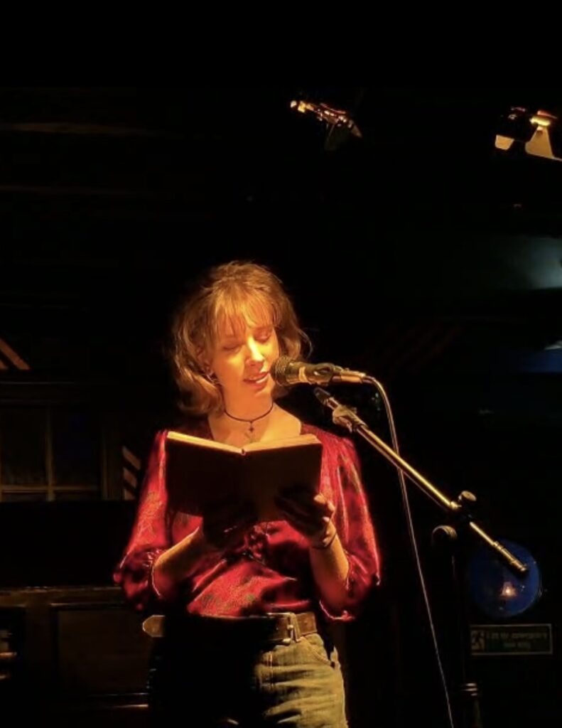 A profile image of Lauren. She is reading from a book into a microphone on a stage. She is brightly lit, whilst her surroundings are darkened.