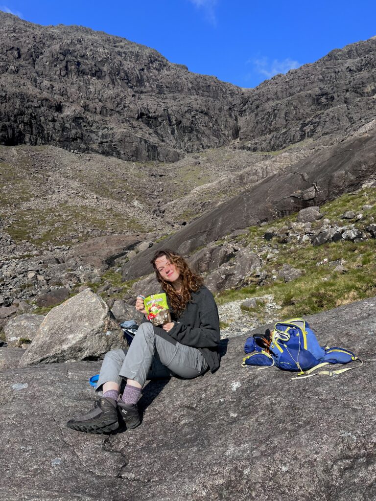 A profile image of Eilidh. She is situated amongst a rocky locale, dressed in hiking gear and eating from a bag labelled 'adventure food'.