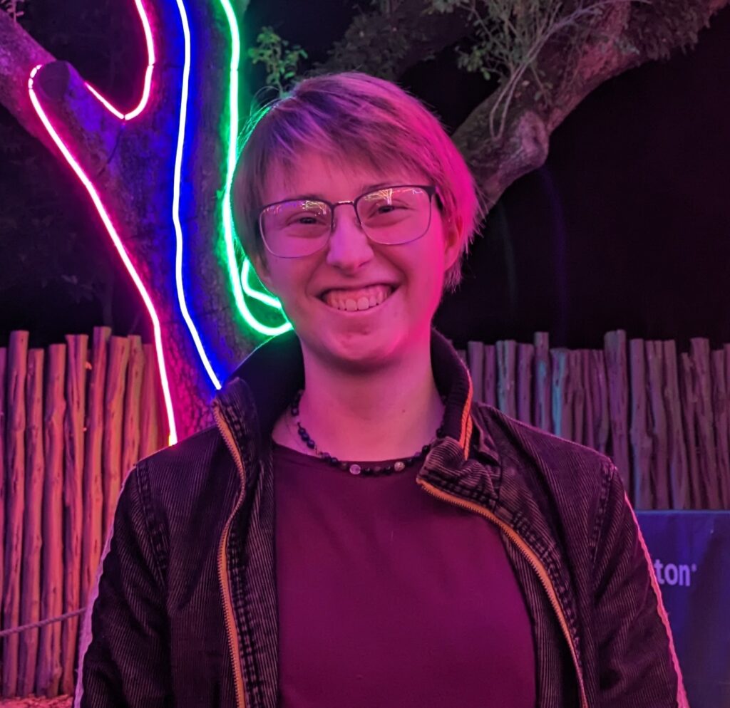 A profile image of Eli. They are smiling and standing in front of a tree that is festooned in neon lighting.