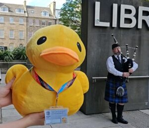 A large soft stuffed duck toy being held next to a piper