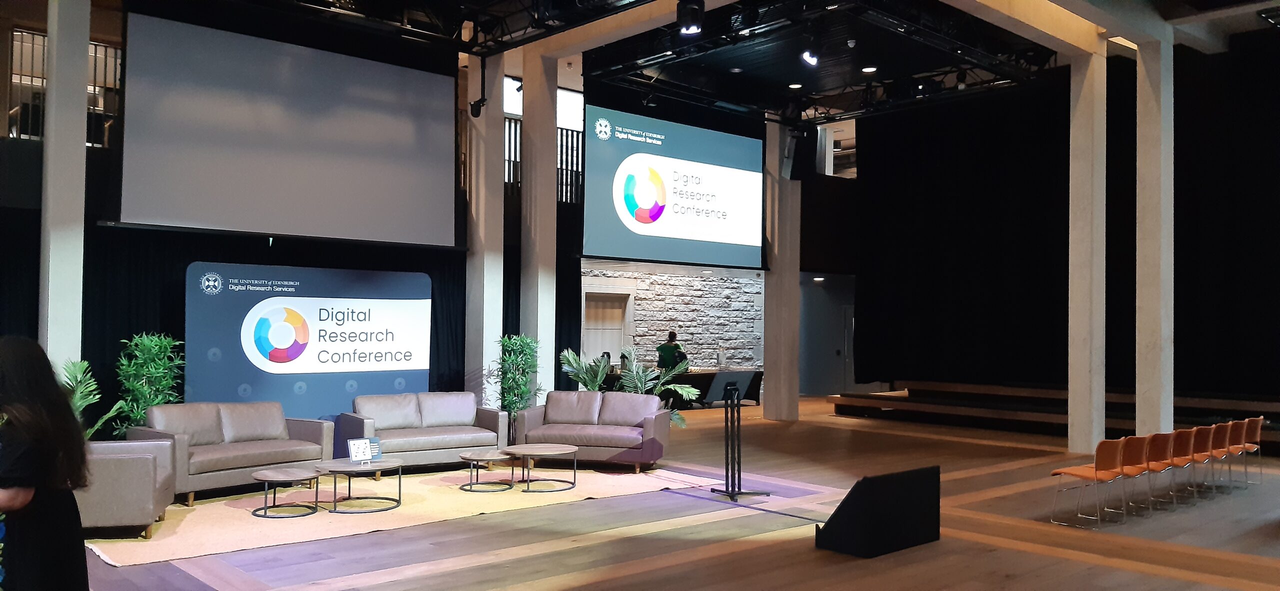 Conference stage set with grey lounges and a large screen above. The screen has an image which says Digital Research Conference. This image is for decorative purposes only