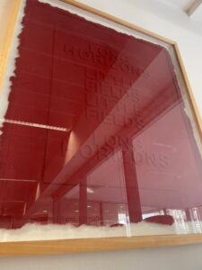 A dark red page of pressed paper pulp reveals text in relief: LONG HORIZONS LITTLE FIELDS LITTLE FIELDS LONG HORIZONS. The page has scraggly edges and is framed and hung on a blank white wall.