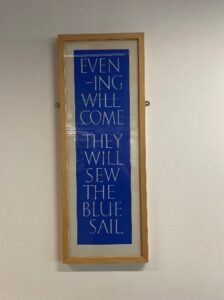 White text on a bright blue background read: EVEN-ING WILL COME THEY WILL SEW THE BLUE SAIL. The page is framed and hung on a plain white wall.