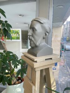 A grey bust of a man with a handlebar moustache sits atop a platform in a sunny corner, beside a potted plant