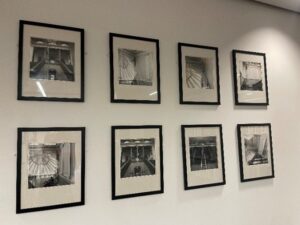 8 framed black and white photographs hang on a white wall, showing a man painting near the ceiling of a Beaux Arts style atrium