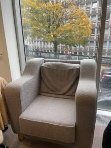 A large gray fuzzy armchair with large thick arms and a squashed cushion, with a window behind it showing Argyle House and a tree with yellow leaves.