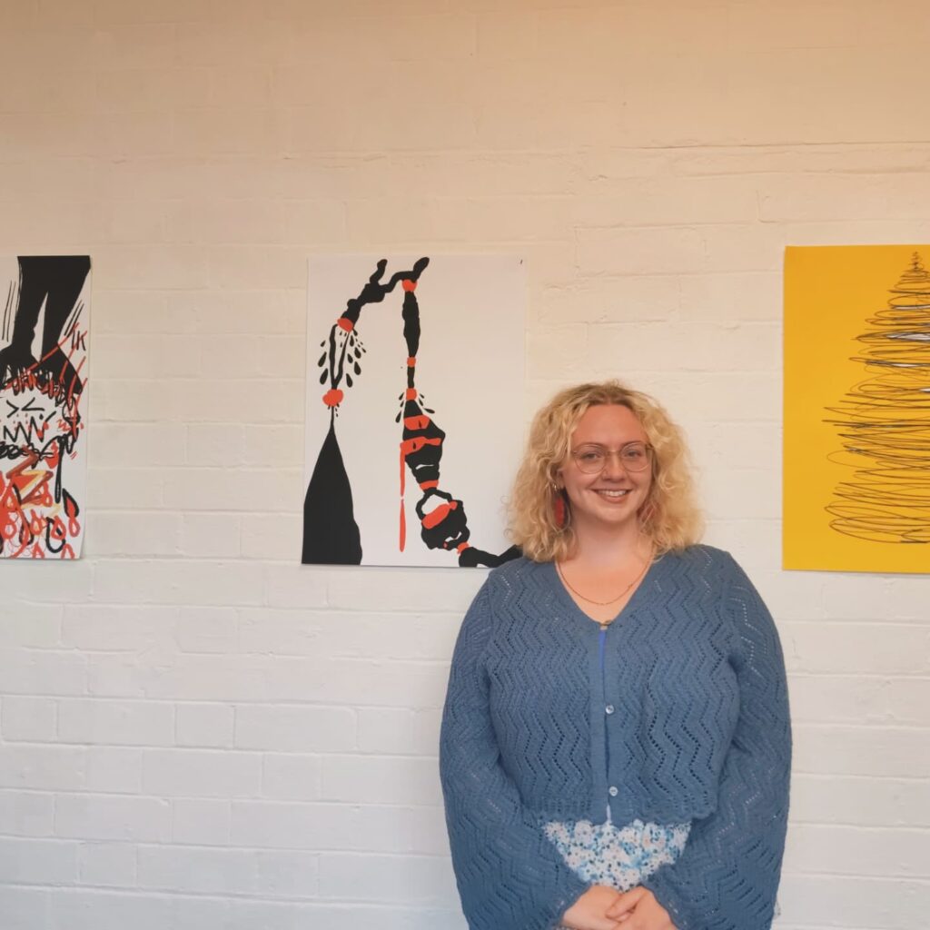 Gemma stands in front of three pieces of her 2D artwork, which are hung on a white wall.