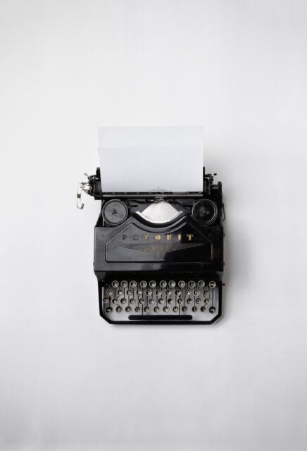 A vintage black type writer with a clean sheet of paper rests on a white table.