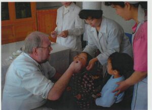 A photograph of Dr. Jim Hoare working as part of a UNICEF and diplomatic team helping with a polio vaccination campaign in Haeju, North Korea. Dr. Hoare is pictured giving the vaccination to a child, surrounded by another child and medical staff. 