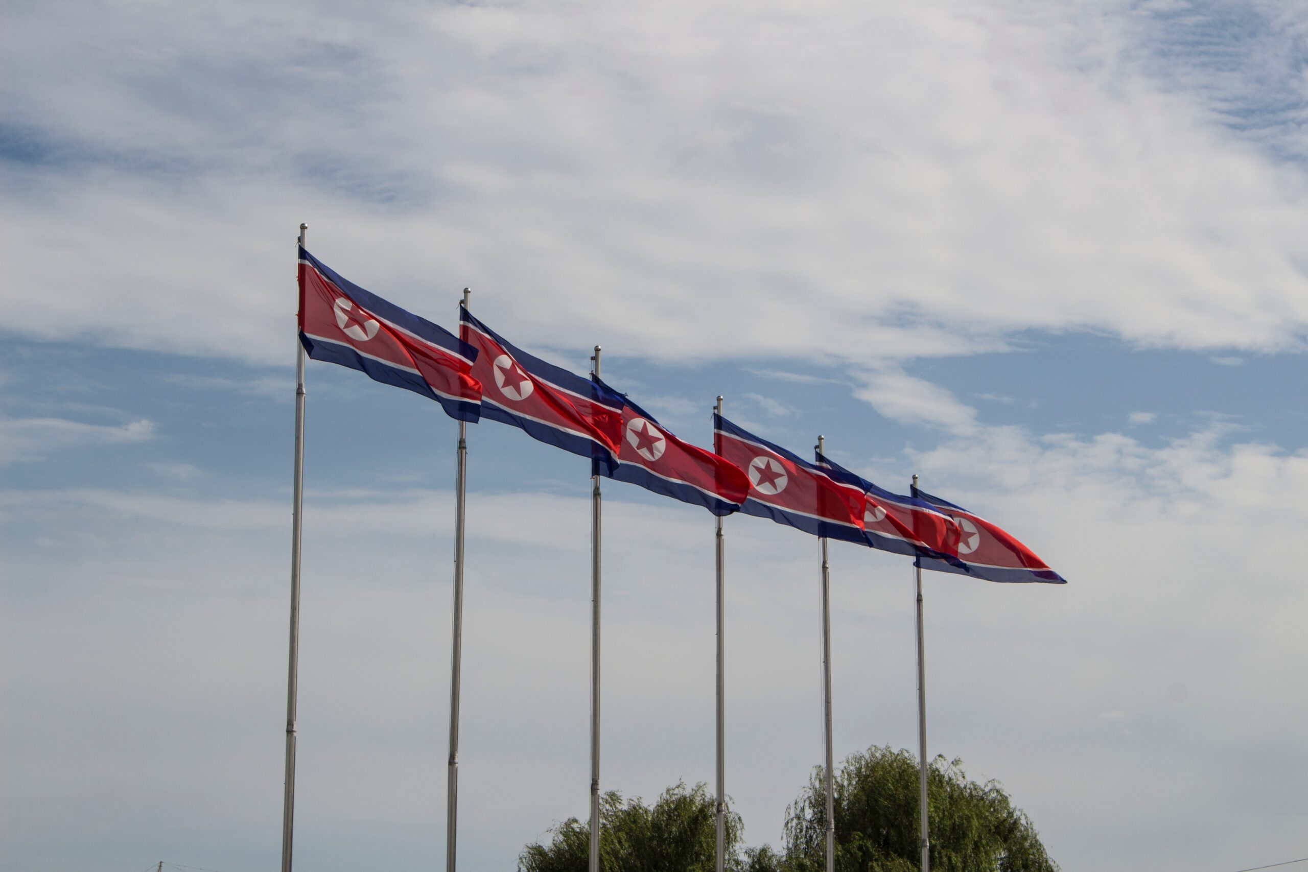 A photograph of a row of flagpoles with flags of the Democratic People's Republic of Korea against a blue sky with clouds.