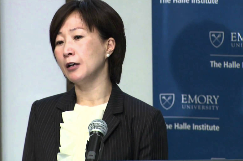A colour photograph of Mikyoung Kim, who has dark hair and is wearing a dark pinstriped suit jacket, a white blouse and earrings, and speaking into a microphone.