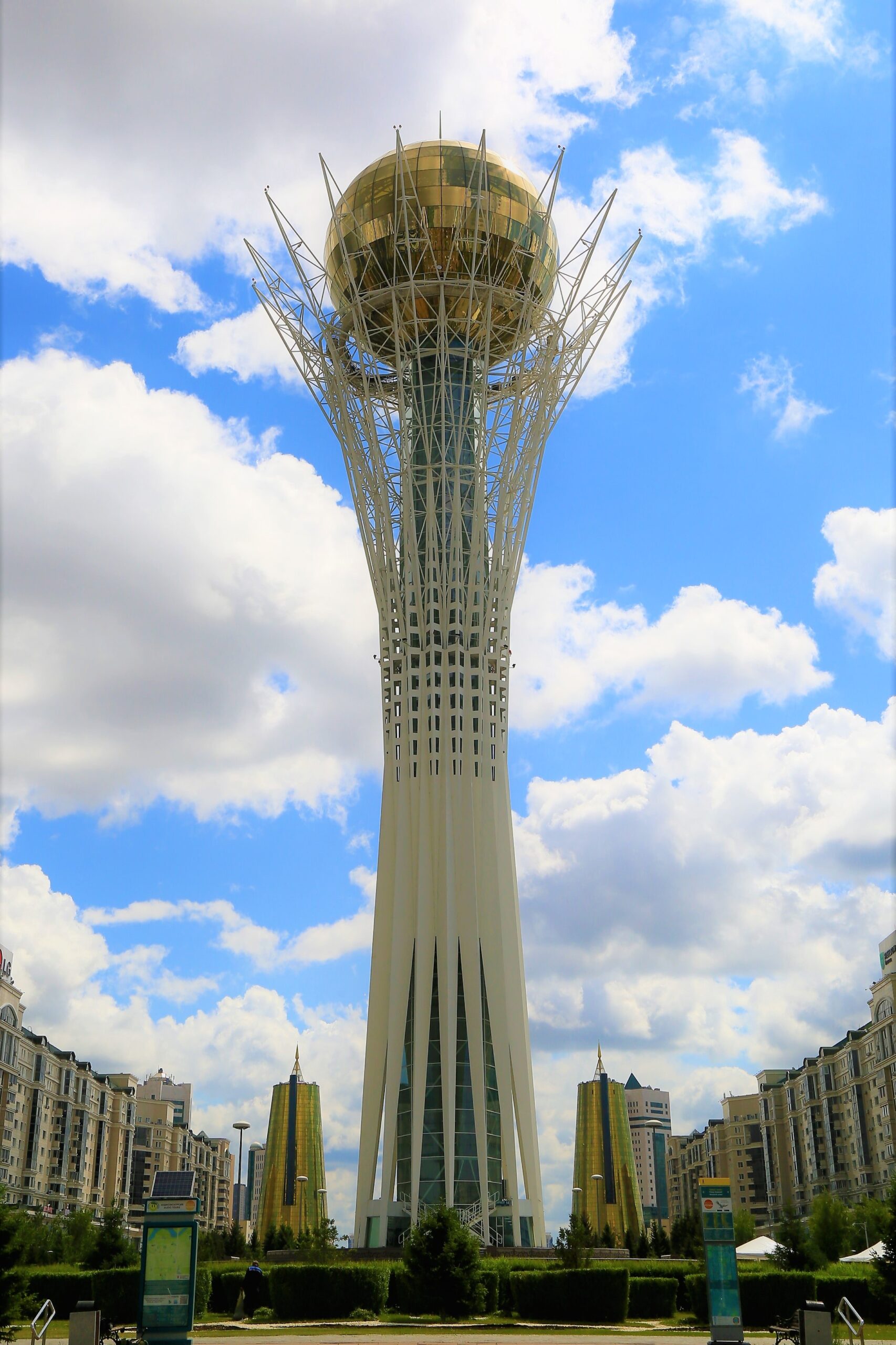 A photograph of the Baiterek monument and observation tower in Nur-Sultan, Kazakhstan. The monument is surrounded by buildings and behind it is a blue sky with clouds.