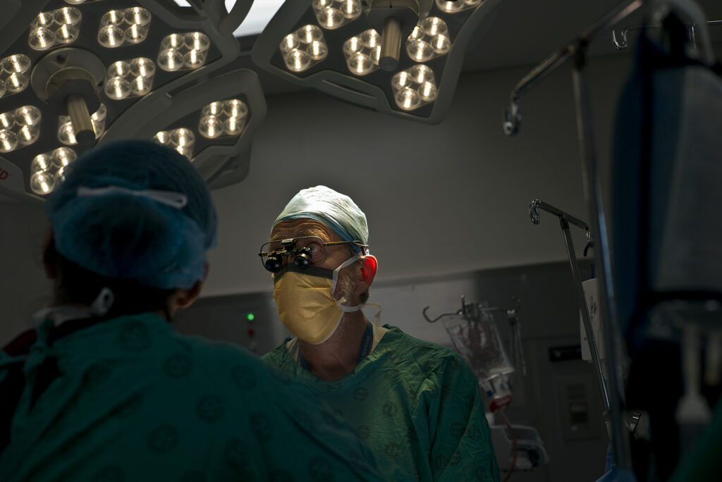 Two surgeons in scrubs, masks, and hats with surgical theatre lights in the background.