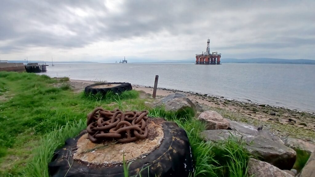 At a beach in Cromarty looking out over a series of oil platforms receding into the distance. In the foreground green grass, old tyres and rusted chains